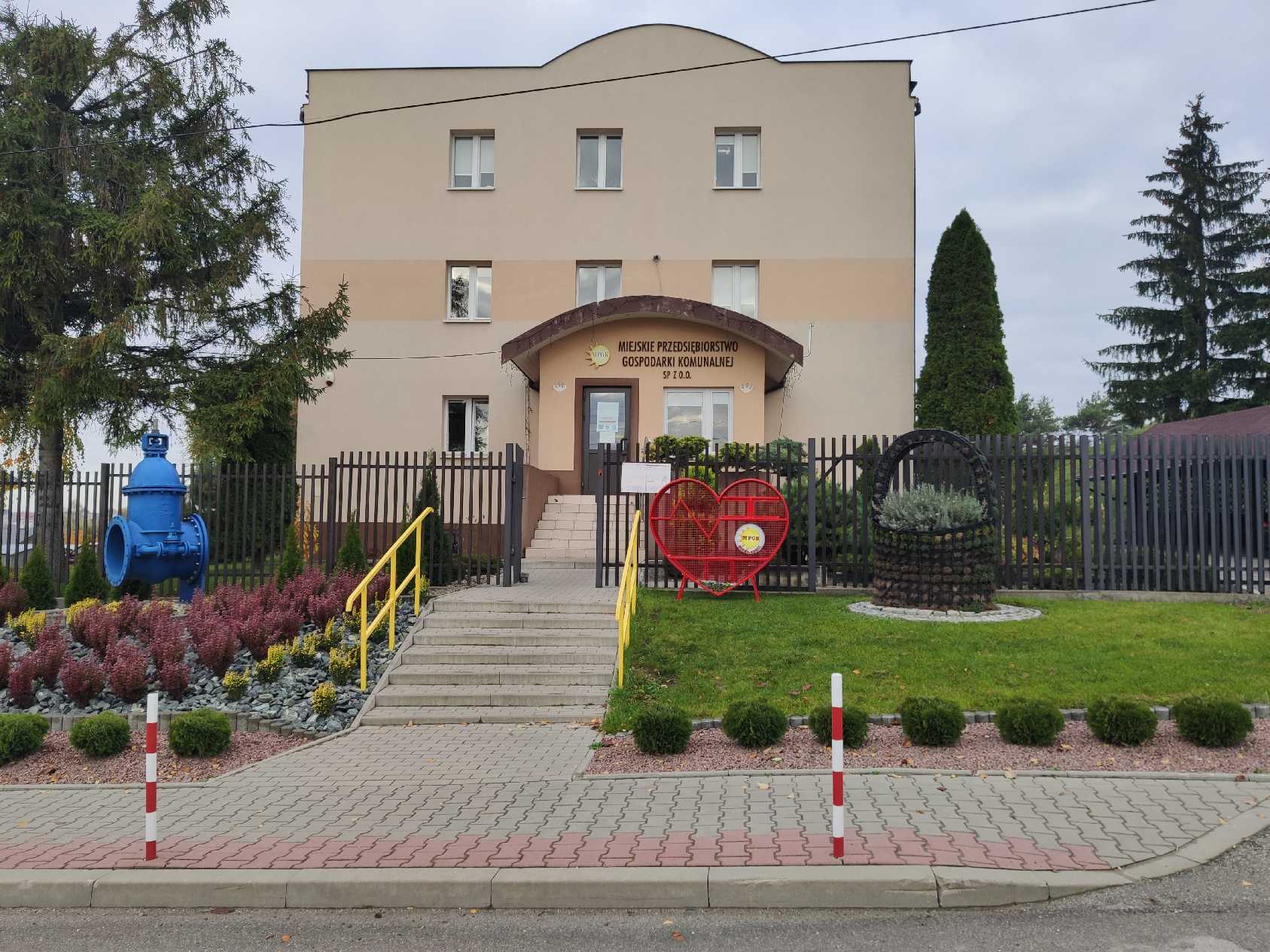 Zdjęcie przedstawia metalowy pojemnik w kształcie serca na nakrętki na tle budynku MPGK w Busku-Zdroju 