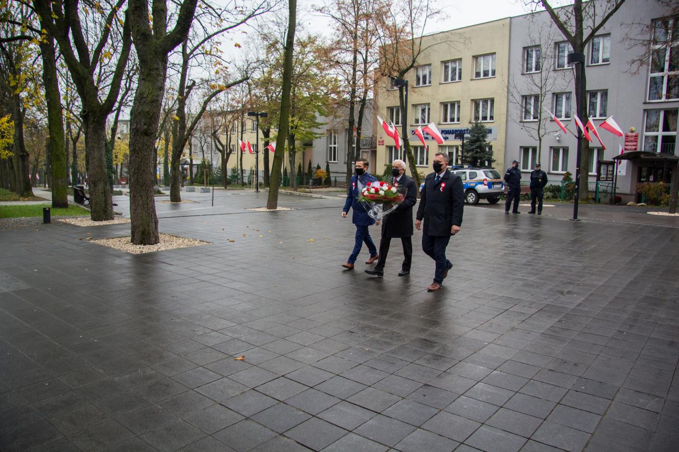 Zdjęcie przedstawia Roberta Burchana Przewodniczącego Rady Miejskiej, Waldemara Sikorę Burmistrza Miasta i Gminy Busko-Zdrój niosącego wiązankę biało-czerwonych kwiatów oraz Michała Marońskiego Z-cę Burmistrza Miasta i Gminy Busko-Zdrój  idących w kierunku pomnika Tadeusza Kościuszki. 