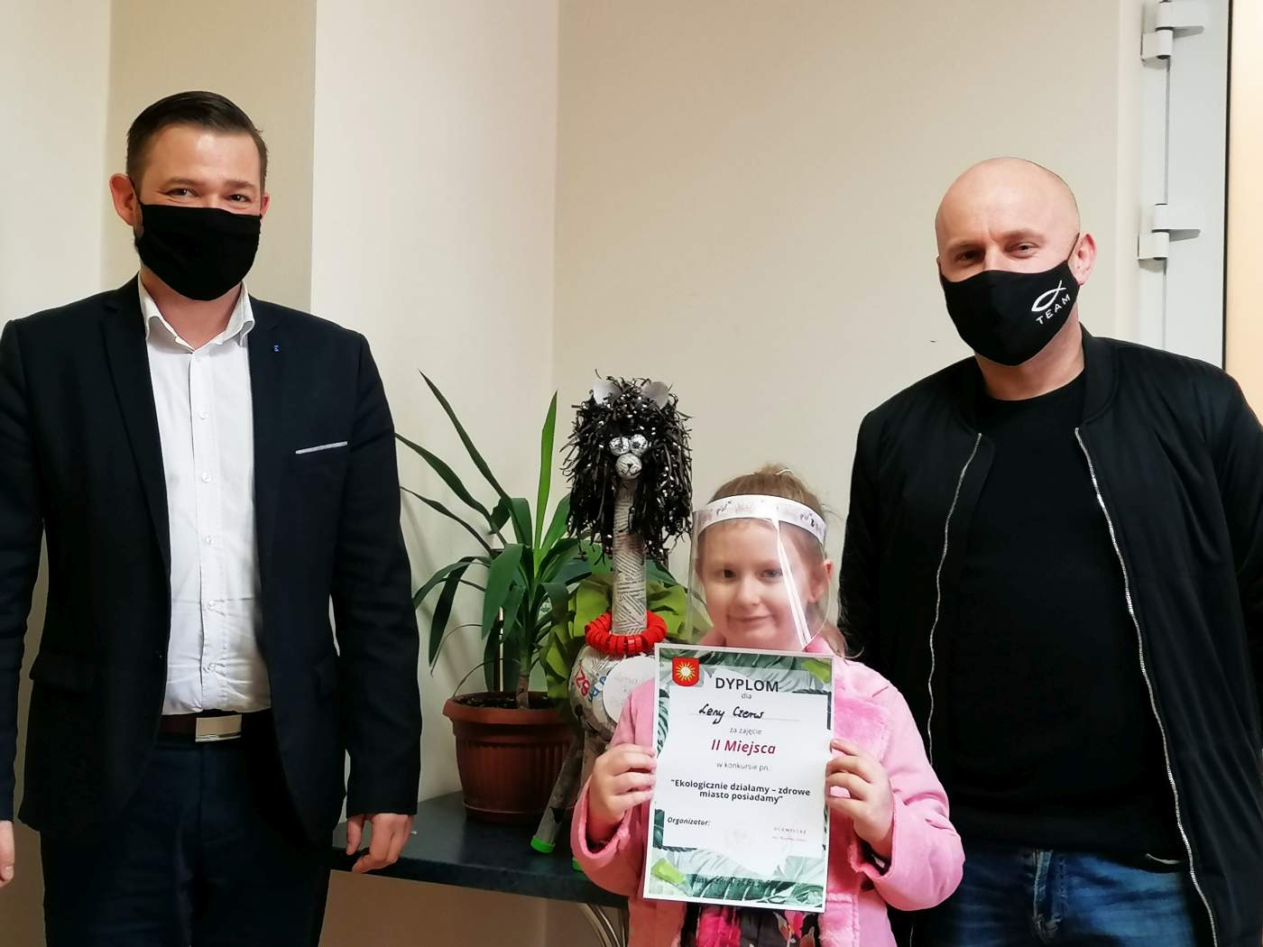 Zdjęcie przedstawia Laureatkę Konkursu trzymającą dyplom wręczony przez Z-cę Burmistrza Miasta i Gminy Michała Marońskiego na tle którego widać nagrodzoną pracę