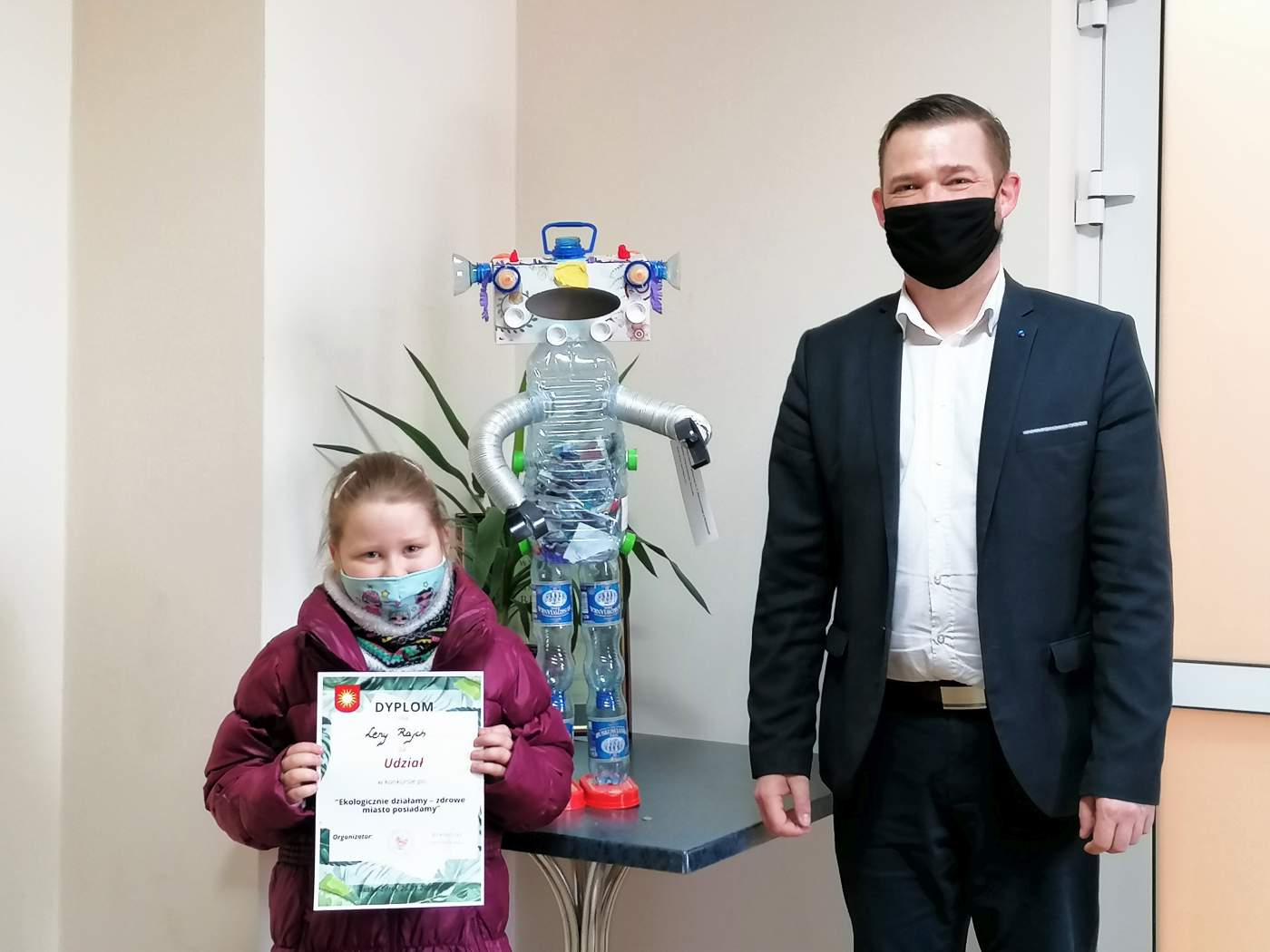 Zdjęcie przedstawia Laureatkę Konkursu trzymającą dyplom wręczony przez Z-cę Burmistrza Miasta i Gminy Michała Marońskiego w tle którego widać nagrodzoną pracę